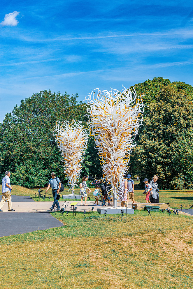 The image depicts a park scene with a large, intricate sculpture made of white and yellow twisted elements. Several people are walking around and observing the sculpture, which is situated on a paved area surrounded by grass and trees under a clear blue sky..