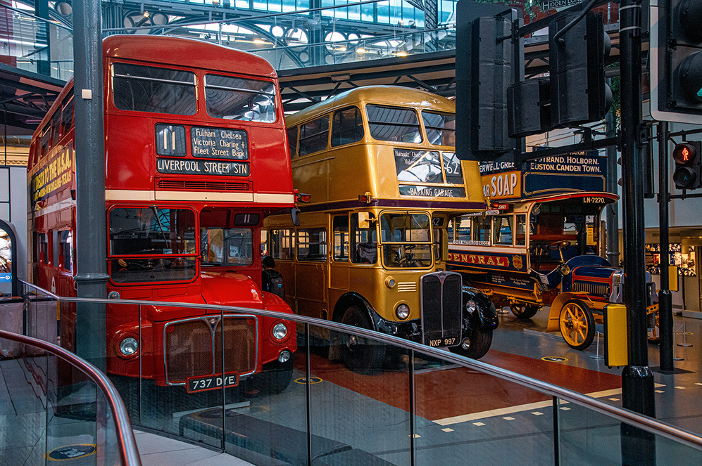 The image depicts a display of vintage double-decker buses in what appears to be a museum or exhibition setting. The buses are painted in distinctive red and yellow colors and are positioned as if they are on a street, complete with traffic lights and street signs. The setting suggests a historical or nostalgic theme, showcasing the evolution of public transportation.