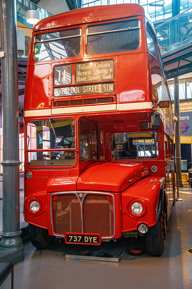 The image depicts a classic red double-decker bus, likely a historical model, displayed indoors. The bus has a route sign indicating destiUKs such as Fulham, Chelsea, Victoria, Charing, Fleet Street, and Bank, with the final stop at Liverpool Street Station. The bus's registration number is 737 DYE. The setting appears to be a museum or an exhibition hall, as suggested by the polished floor and the presence of other exhibits in the background