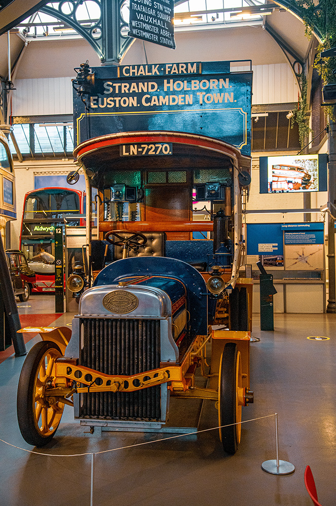 The image depicts a vintage bus on display in a museum. The bus has a sign on top indicating routes to Chalk Farm, Strand, Holborn, Euston, and Camden Town. The bus is marked with the identification number LN-7270. The display is part of a larger exhibit, with other vintage vehicles and informational plaques visible in the background.