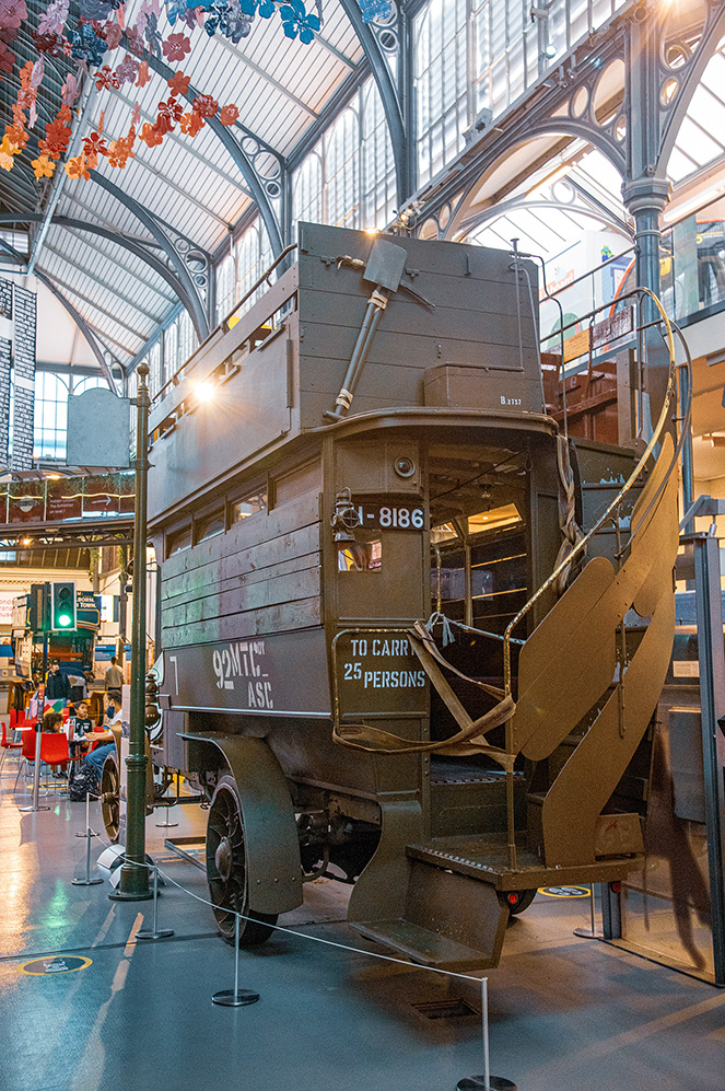 The image depicts a vintage tram or bus on display in a museum-like setting with a high, arched glass ceiling and decorative elements. The vehicle is marked with the number 8186 and has a capacity of 25 persons. The interior of the vehicle is visible, showing wooden benches. The area around the vehicle is cordoned off with stanchions and ropes, indicating it is part of an exhibit. The setting suggests a historical or transportation museum.