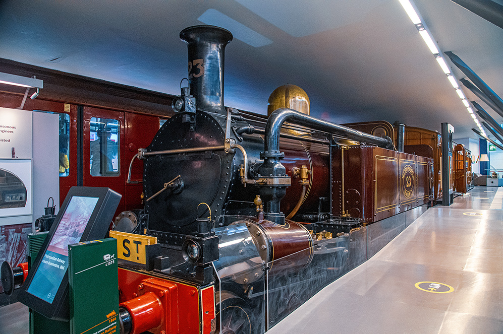 The image depicts a vintage steam locomotive on display in a museum. The locomotive, labeled with the number 23, is prominently featured with its intricate details and polished exterior. The display includes informational screens and signs, providing visitors with historical context and information about the exhibit. The setting appears to be well-lit and spacious, with other exhibits visible in the background.