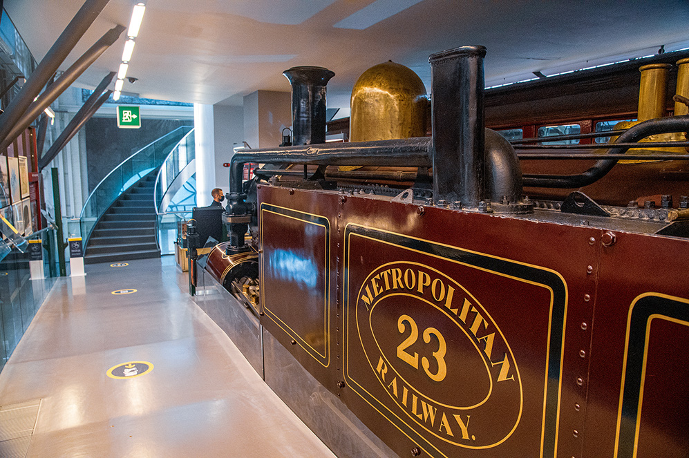 The image shows a vintage steam locomotive labeled 'Metropolitan Railway 23' on display in a modern, well-lit museum setting. The locomotive is polished and well-maintained, with a prominent gold and black color scheme. The museum features a staircase and an escalator in the background, indicating a multi-level exhibit space. The setting suggests an environment dedicated to preserving and showcasing historical railway artifacts.