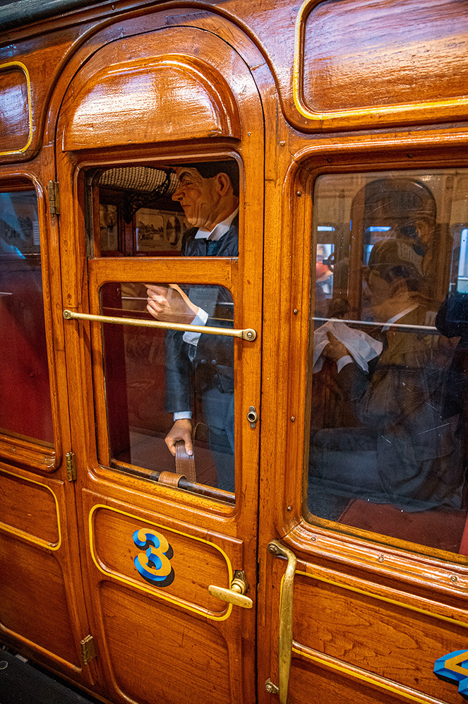The image depicts a vintage train compartment with wooden interiors and large windows. Inside, there are two wax figures dressed in early 20th-century attire, with one figure leaning out of the open window and the other seated inside. The door of the compartment is marked with the number 3 and features brass fittings. The overall scene suggests a historical or museum setting.