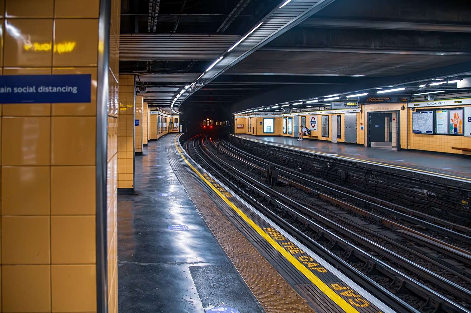 The image depicts an empty subway station with a curved platform and tracks extending into a dark tunnel. The station is well-lit with overhead lights, and there is a sign on the left wall reminding passengers to maintain social distancing. The platform has yellow safety lines and tactile paving for visually impaired individuals. The station appears clean and well-maintained, with various signs and advertisements on the walls.