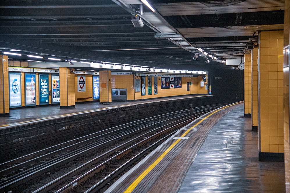 The image depicts an empty subway station with yellow-tiled walls, tracks in the center, and various advertisements on the platforms. The station appears clean and well-lit, with signs indicating it is the Cannon_Street_Station.