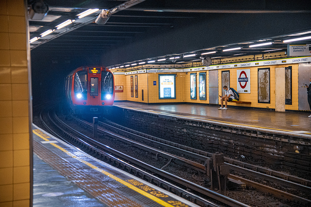 The image depicts a subway station with a train arriving at the platform. The station is identified as Cannon Street, part of the London Underground system. The platform is relatively empty, with only a few people visible. The station's design includes yellow tiles and typical underground signage. The train is red, indicating it is part of the London Underground network.
