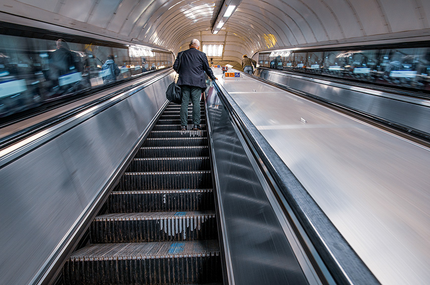 The image depicts a man standing on an escalator in a subway station, moving upwards. The station appears to be clean and well-lit, with other people visible in the background, some of whom are also on escalators. The man is wearing a dark jacket and green pants, and he is carrying a bag. The escalator is made of metal with visible steps and side panels.