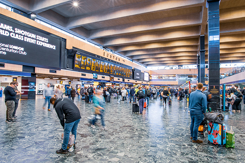 The image depicts a busy train station with numerous people moving about, some carrying luggage. The station has a large, modern interior with high ceilings and a tiled floor. There are digital departure and arrival boards prominently displayed in the background. Some individuals appear to be bending down, possibly tying their shoes or picking something up. The overall atmosphere suggests a typical bustling scene at a major transportation hub
