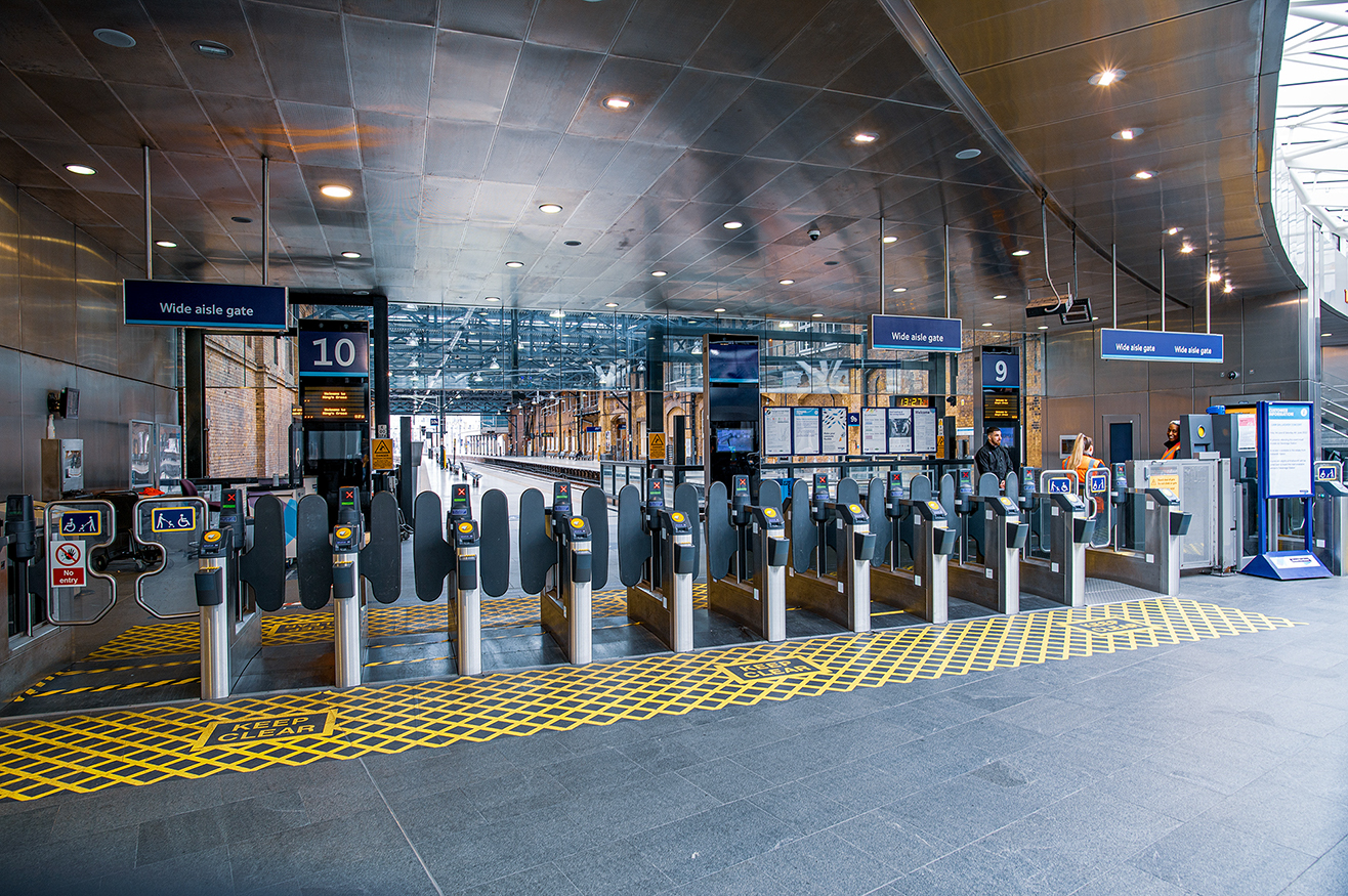 The image shows a modern train station entrance with multiple automated turnstile gates labeled as 'Wide aisle gate'. There are digital displays above the gates, showing information and time. The station has clear signage, a clean environment, and a few people are seen entering through the gates. The floor has yellow tactile paving for assisting visually impaired individuals, and there are various signs and posters on the walls.