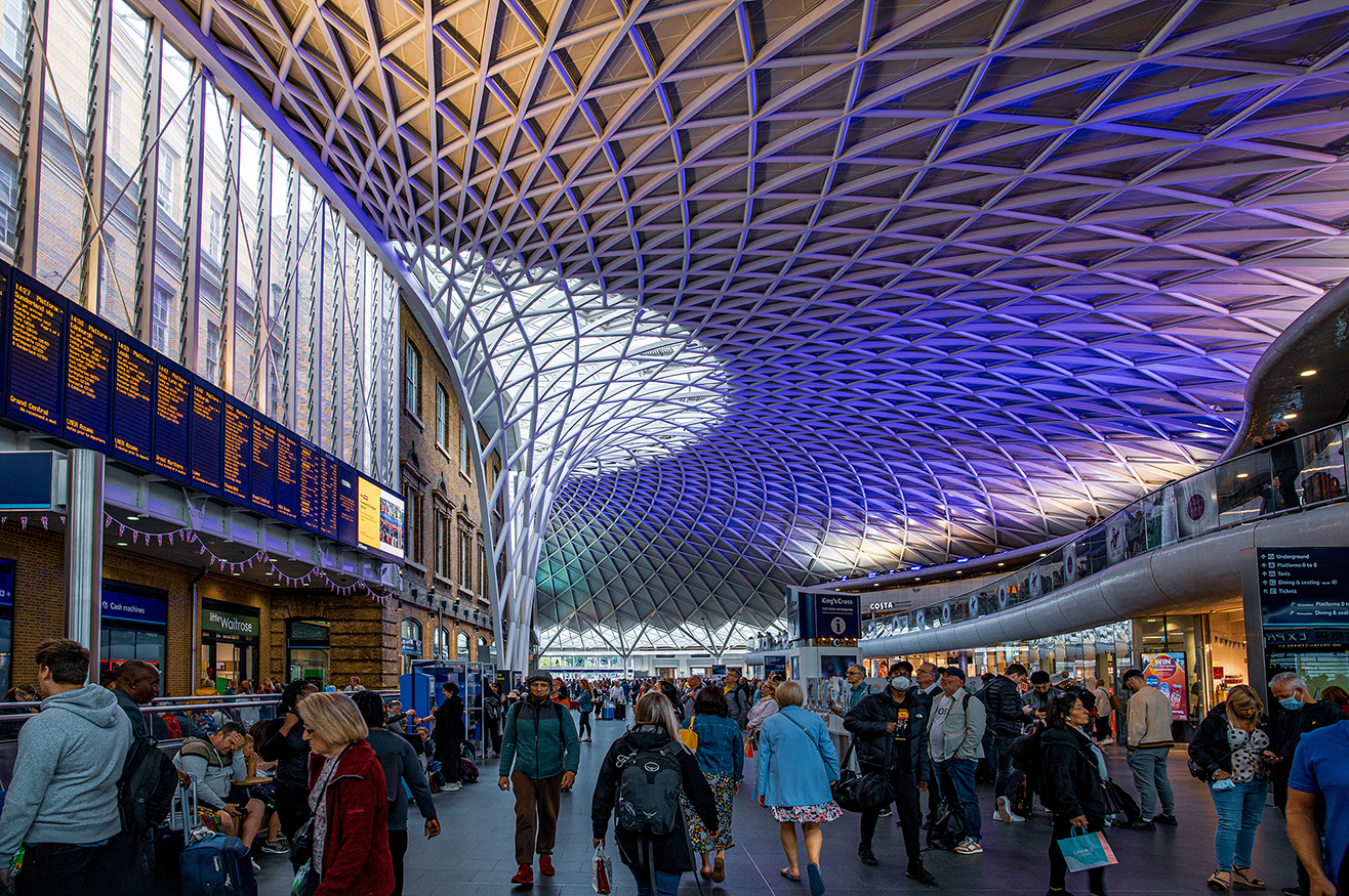 The image depicts a bustling train station with a modern, architecturally striking roof made of glass and steel. The station is filled with people, some walking and others waiting. There are multiple digital display boards showing train schedules and other information. Various shops and facilities, such as WHSmith and Costa Coffee, are visible along the sides of the station. The overall atmosphere is busy and vibrant, indicative of a major transportation hub.