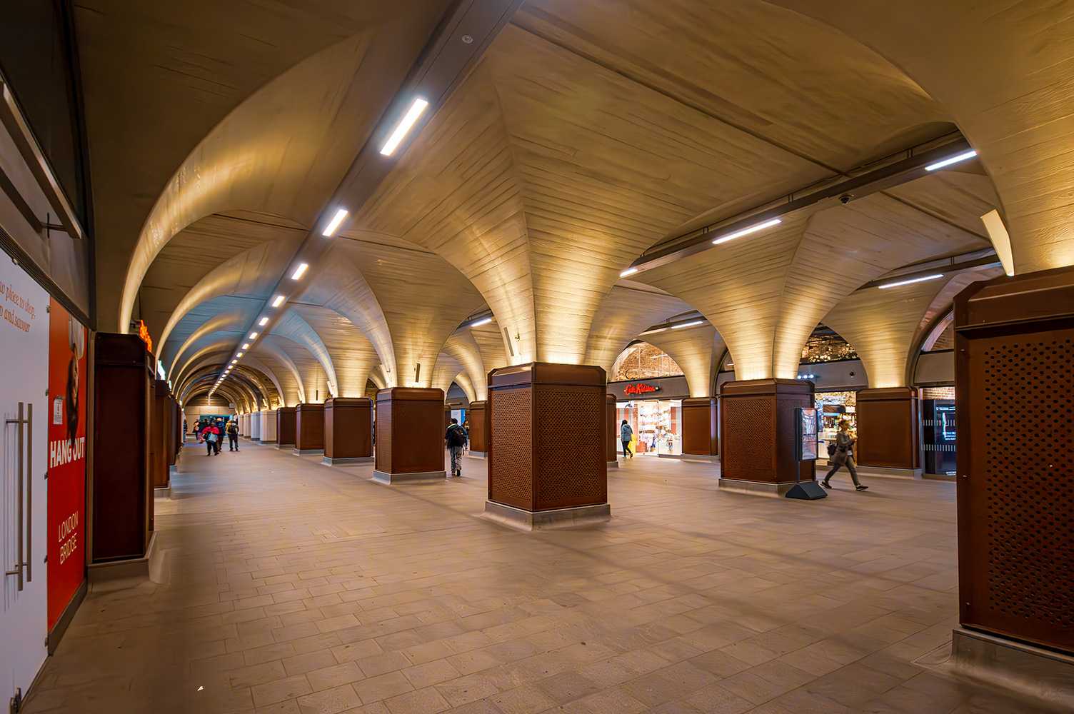 The image depicts an indoor shopping arcade with a series of arched ceilings lined with wooden panels and illuminated by linear lights. The floor is tiled, and there are several large, square pillars with textured surfaces. The arcade features various shops, including one with a prominent red sign that reads 'HAY.' The space is relatively empty, with only a few people visible in the distance, suggesting a quiet moment in the shopping area.