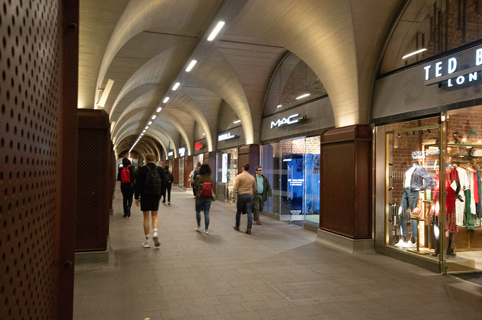 The image depicts an indoor shopping arcade with arched ceilings and various storefronts. People are walking through the corridor, and there are visible signs for stores such as MAC and Ted Baker London. The arcade is well-lit with modern lighting fixtures, and the overall atmosphere appears to be bustling yet orderly.
