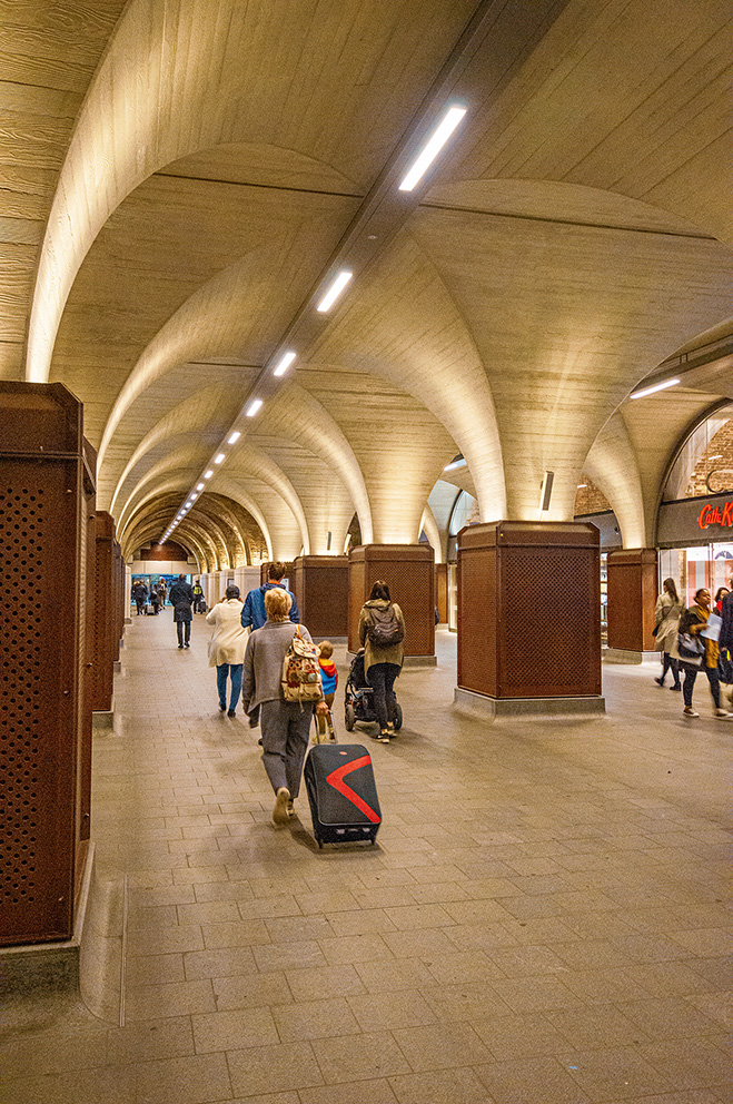 The image depicts a bustling underground passageway with arched ceilings and modern lighting. People are seen walking in both directions, some pulling luggage, indicating it might be a subway or metro station. The walls are adorned with wooden panels, and there are various shops and kiosks along the sides. The overall atmosphere is lively, with individuals engaged in their daily routines.