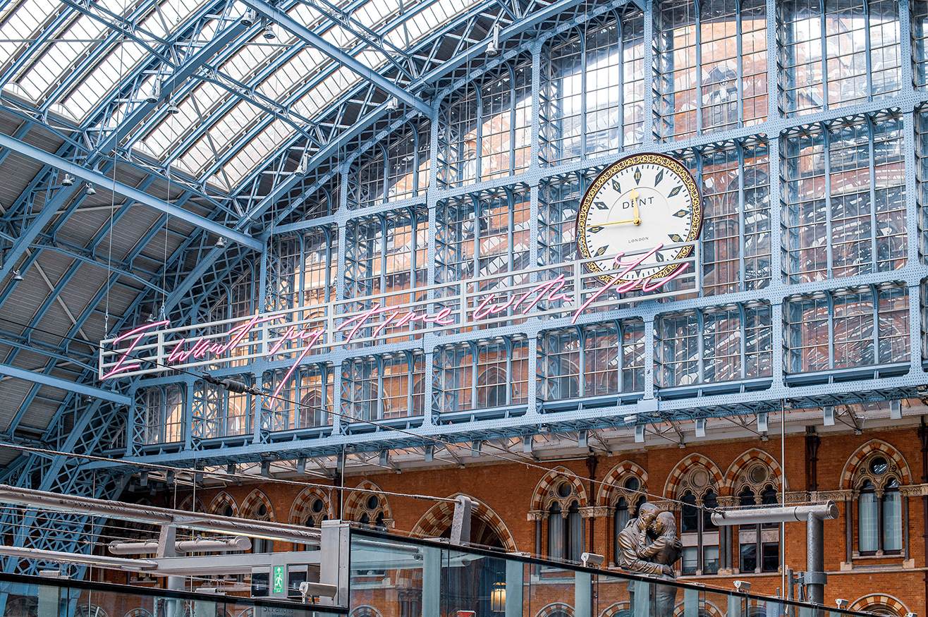The image depicts the interior of a large, historic train station with a high, arched glass roof supported by a steel framework. A prominent clock with the name 'DENT' is centrally located on an upper level. The station features intricate architectural details, including arched windows and decorative elements. There are multiple levels with walkways and railings, and a statue of a man is visible on the lower level. The station appears to be bustling, with signs indicating directions and exits.