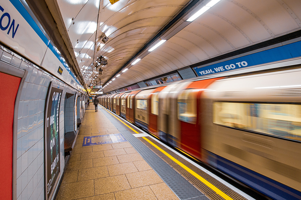 he image depicts a subway station with a train arriving or departing. The station features tiled walls with advertisements, a platform marked with safety tiles, and overhead lighting. The train is in motion, creating a sense of speed. The station appears clean and well-maintained, with clear signage and a relatively empty platform.