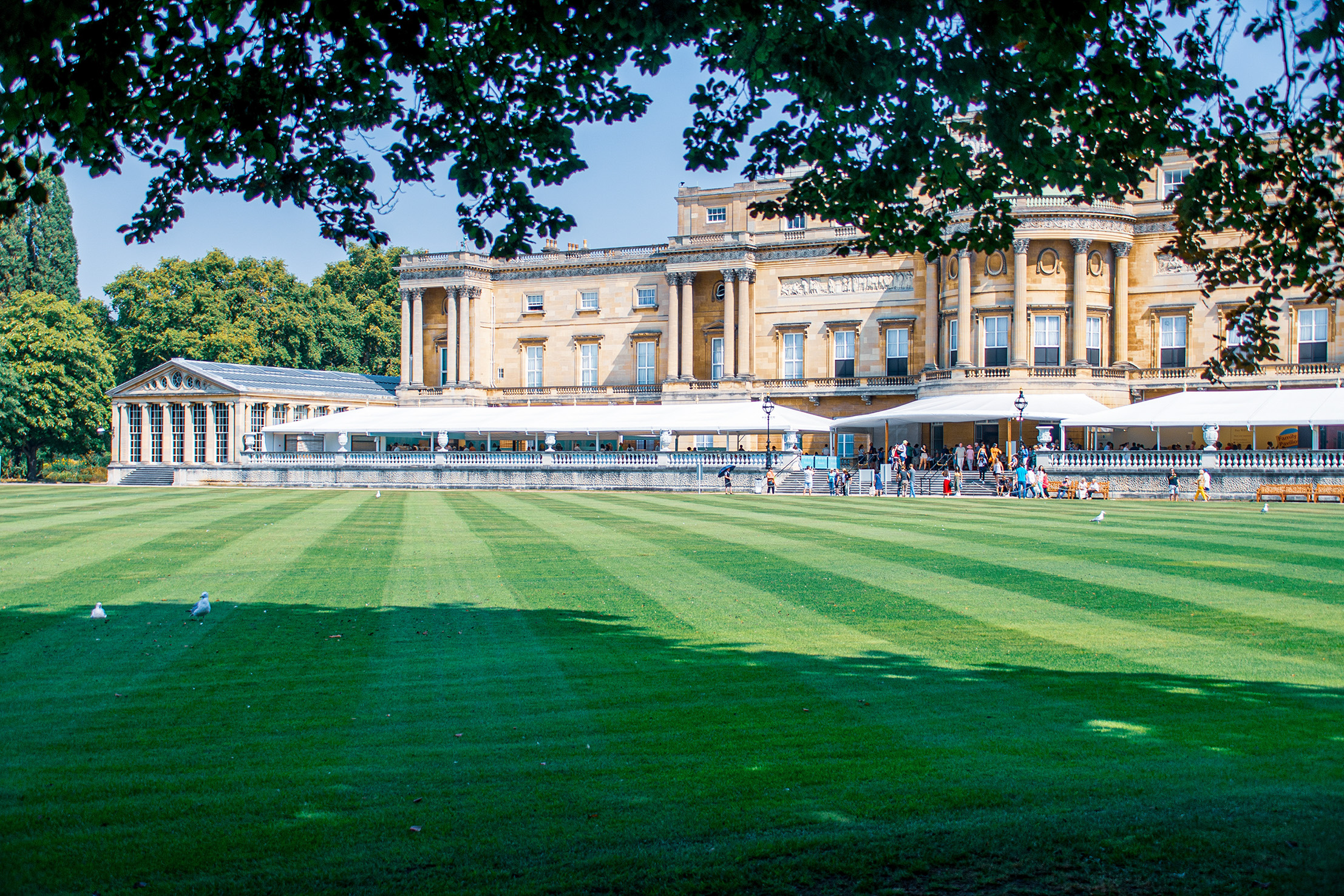 The image depicts a grand, historic building with classical architecture, likely a palace or mansion, surrounded by a well-maintained lawn and trees. The building features large windows, columns, and intricate stonework. There are people walking and sitting on the lawn, and several white umbrellas and tables are set up, suggesting an outdoor cafe or seating area. The scene is bright and sunny, indicating a pleasant day.