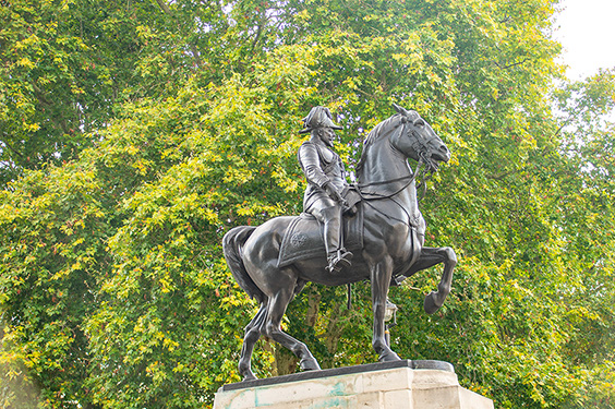 The image depicts an equestrian statue, which is a statue of a person mounted on a horse. The statue is likely made of bronze or a similar material and is mounted on a stone pedestal. The person on the horse appears to be dressed in historical military attire, suggesting that the statue commemorates a historical figure, possibly a military leader or a notable figure from history. The statue is set against a backdrop of lush green trees, indicating that it is located in an outdoor setting, such as a park or a public square.