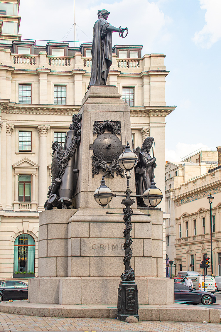 The image depicts a monument with a statue of a figure holding a crown, standing on a pedestal adorned with intricate sculptures and the word 'CRIME' inscribed on it. The pedestal also features multiple lamps and is situated in an urban setting with historic buildings in the background.