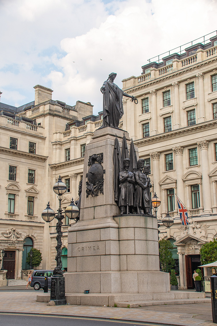 The image depicts a statue in a city setting, likely a monument dedicated to the Crimean War, as indicated by the word 'CRIMEA' on the pedestal. The statue features a central figure holding a scroll and is surrounded by smaller figures. The background shows historic buildings with classical architecture, and a British flag is visible, suggesting the james might be in the United Kingdom.