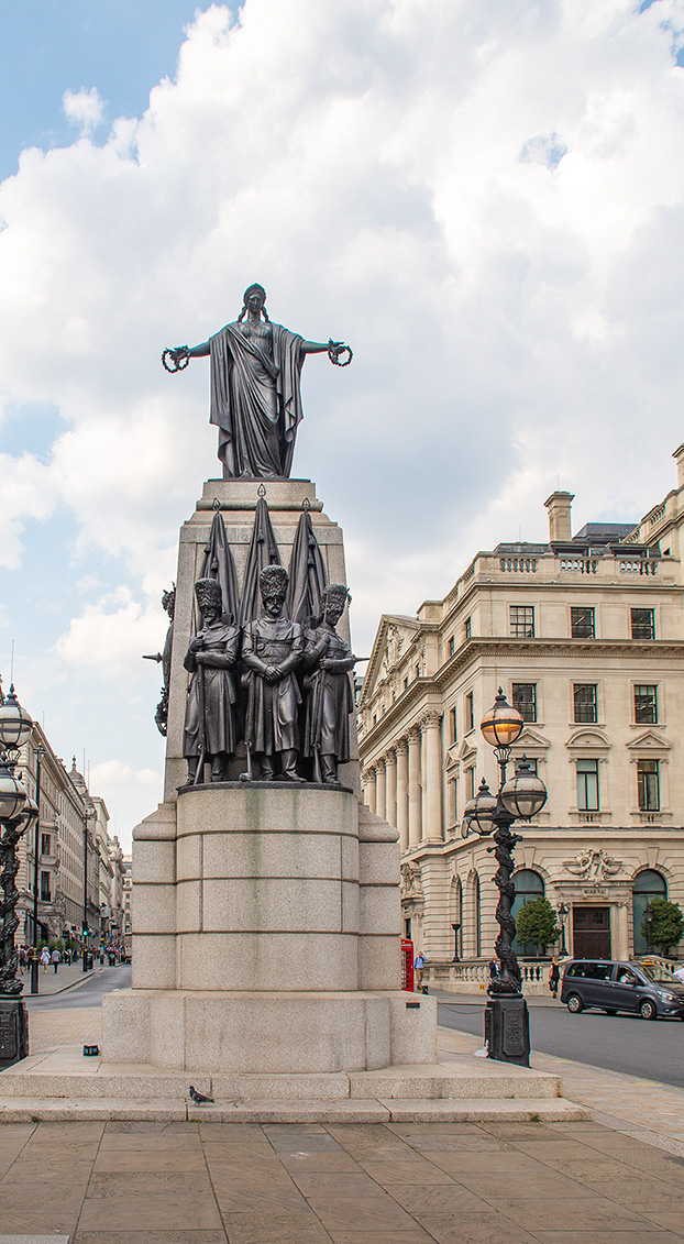 The image depicts a large statue in a city square, featuring a central figure with outstretched arms standing atop a pedestal. Below the central figure, there are four smaller statues grouped together. The statue is surrounded by an ornate lamp post and is situated in front of a grand building with classical architectural features. The scene is set on a clear day with a partly cloudy sky.