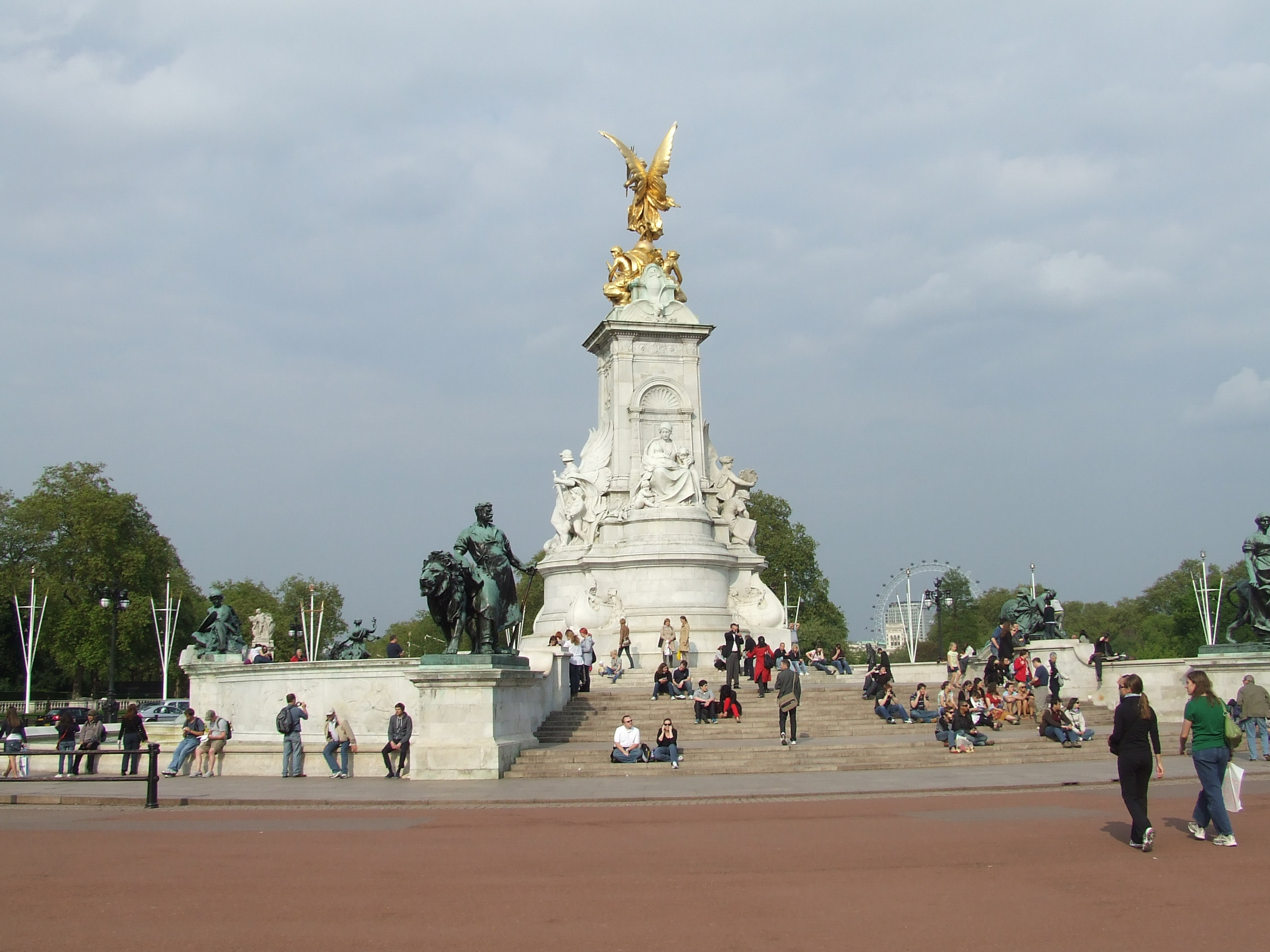 The image depicts the Victoria Memorial in front of Buckingham Palace in London, UK. The memorial features a large white stone structure adorned with statues and a golden winged figure at the top. People are seen sitting on the steps and around the memorial, enjoying the surroundings. The scene is set on a clear day with a partly cloudy sky, and the iconic London Eye Ferris wheel is visible in the background.