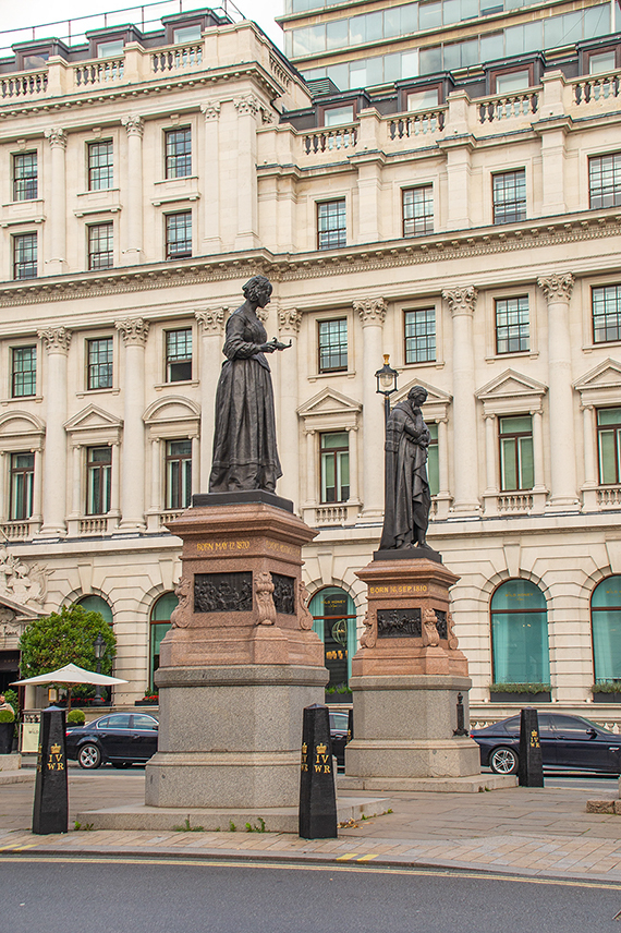 The image depicts a street scene with two statues in front of a large, ornate building. The statues are mounted on substantial pedestals, and the building behind them has classical architectural features, including columns, pilasters, and decorative moldings. The street in front of the building has several cars parked along it, and there is a sidewalk with bollards separating the statues from the roadway.