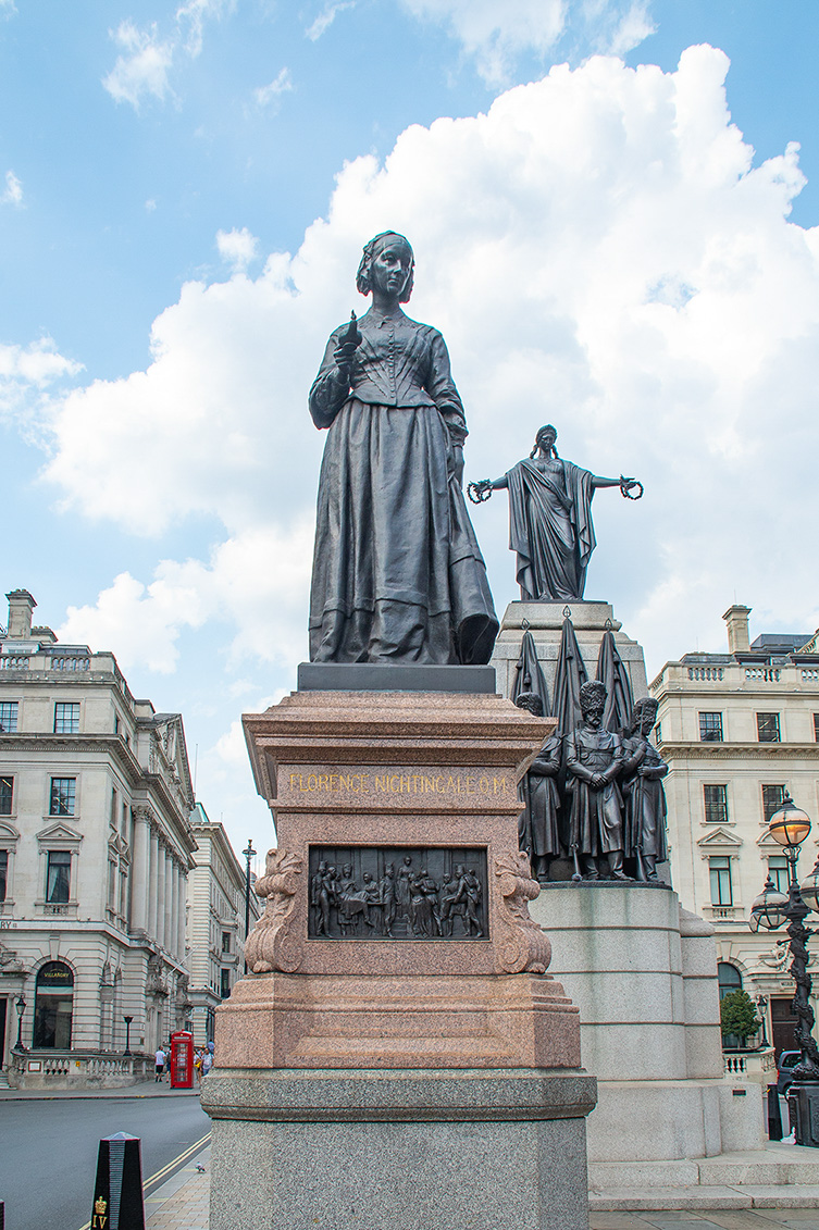 The image depicts a statue of Florence Nightingale, a prominent figure in nursing history, standing prominently in the center. The statue is mounted on a large pedestal with inscriptions and additional sculptural elements around it. The setting appears to be an urban area with classical buildings in the background, suggesting a public square or plaza.