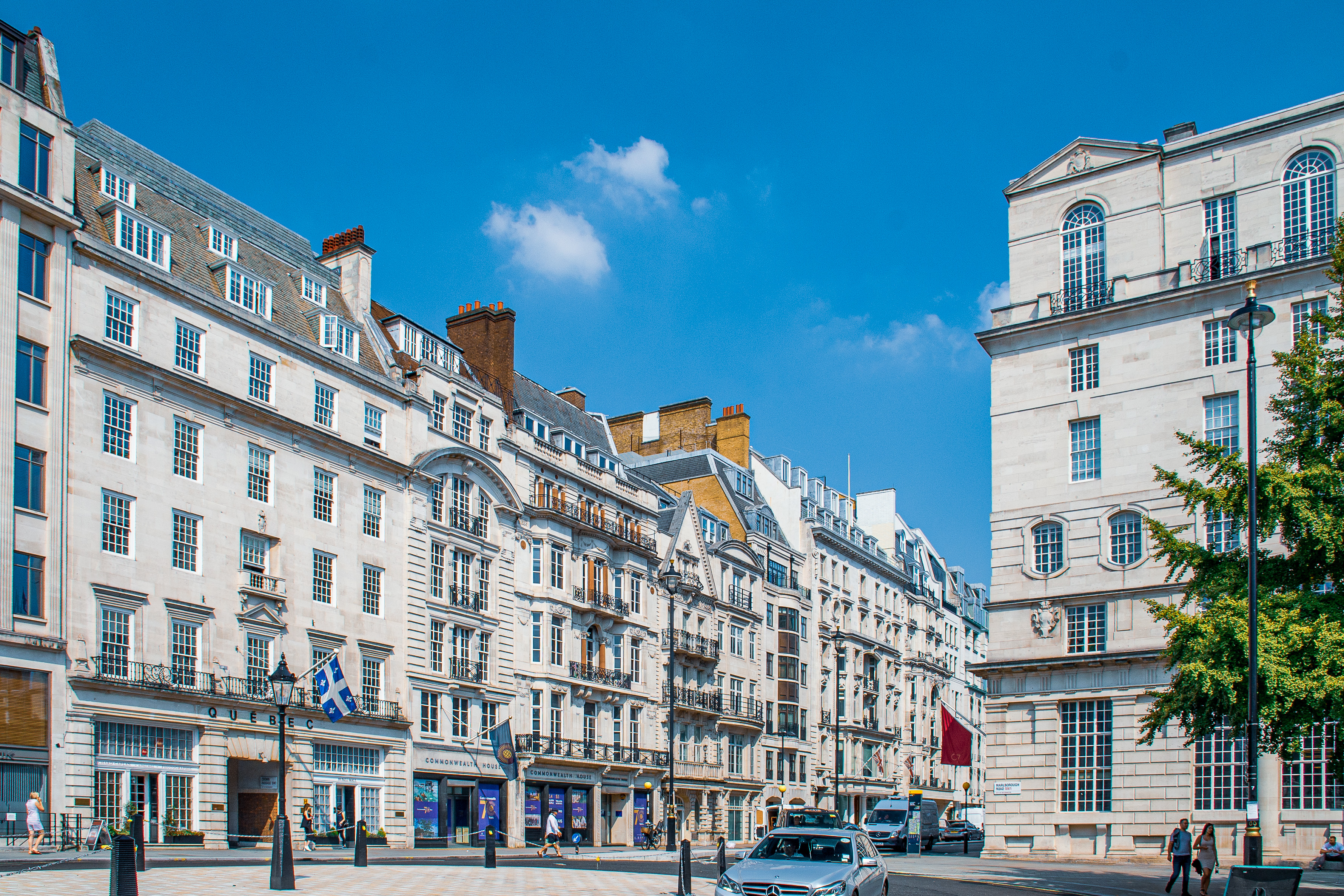 The image depicts a historic brick building with a courtyard. The architecture features large white-framed windows, arched doorways, and a crenelated roofline. The building has multiple chimneys and is constructed from red bricks, giving it a classic and sturdy appearance. The courtyard is paved with red tiles and has some greenery along the edges. The overall scene suggests a well-preserved or restored historical structure, possibly a castle or a manor.