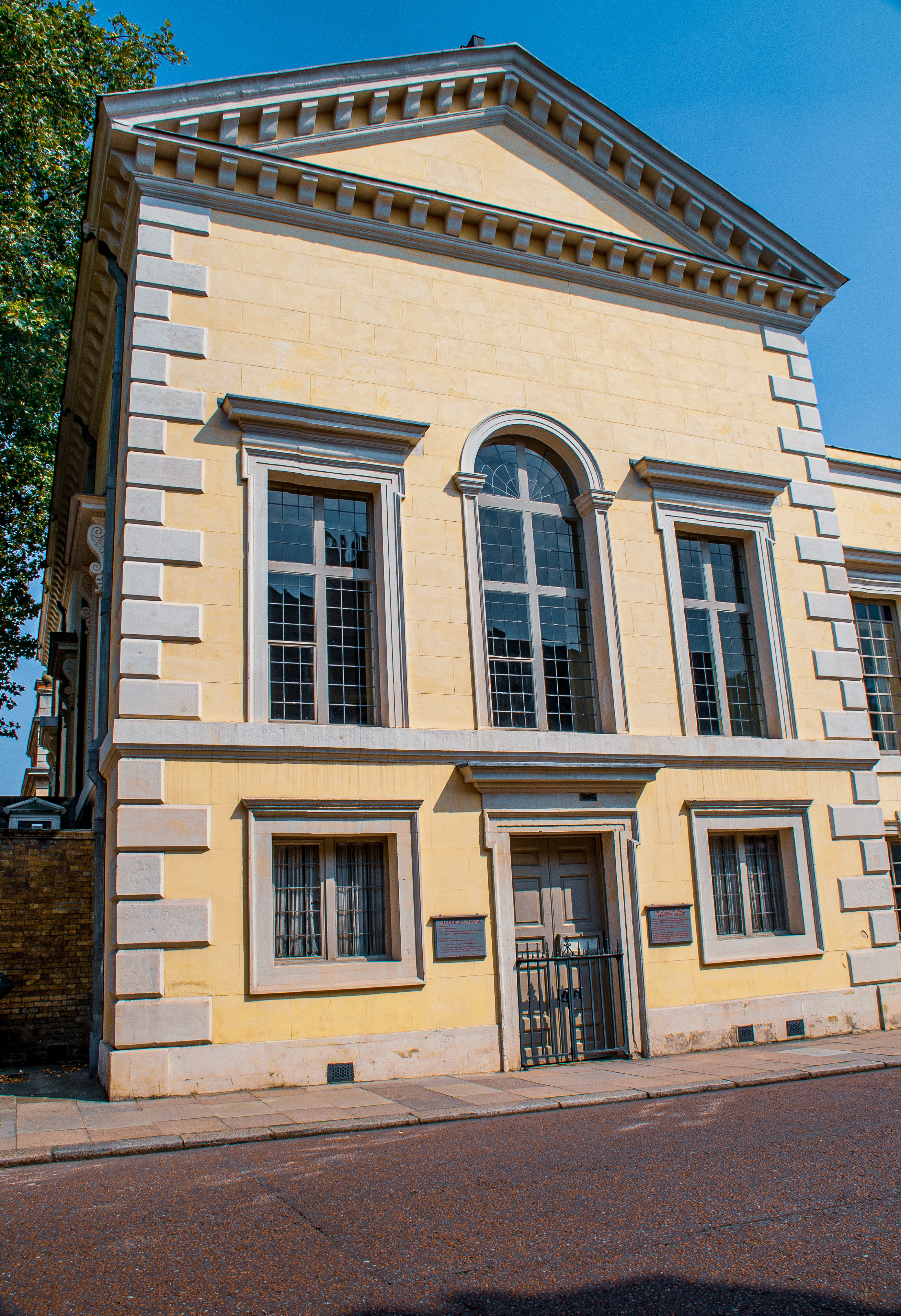 The image depicts a classical-style building with a symmetrical facade, featuring large windows and a prominent triangular pediment. The structure is constructed with light-colored stone blocks and has decorative moldings around the windows and along the roofline. There are two plaques on either side of the entrance, suggesting the building may have historical or institutional significance