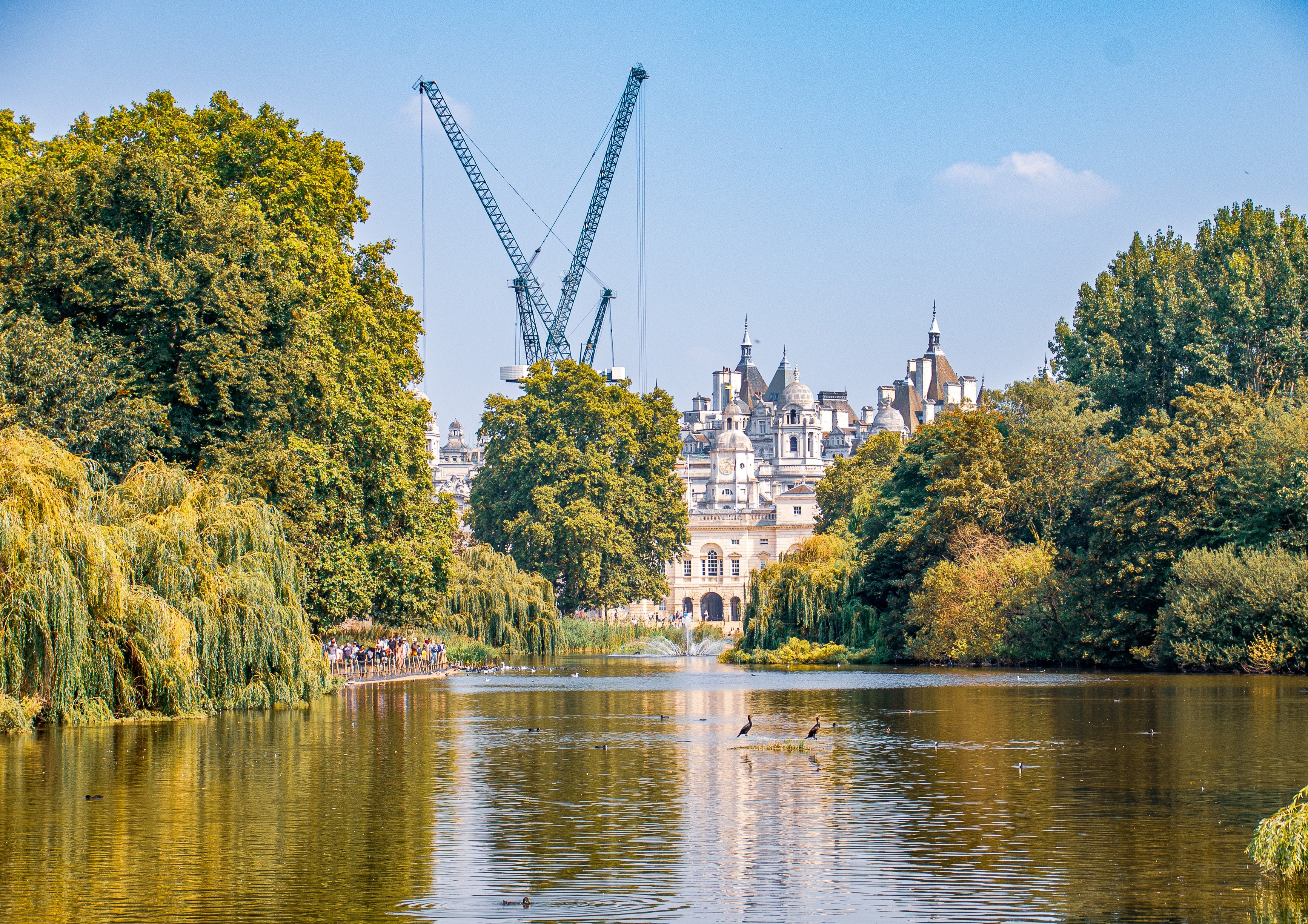 The image depicts a serene park scene with a lake in the foreground, surrounded by lush greenery and trees. In the background, there is an ornate building with multiple spires and domes, partially obscured by the trees. Two large construction cranes are visible behind the building, indicating ongoing construction or renovation work. The sky is clear with a few clouds, suggesting a bright and sunny day. Several birds are seen on the water, and a group of people is gathered near the shore, possibly enjoying a leisurely day in the park.