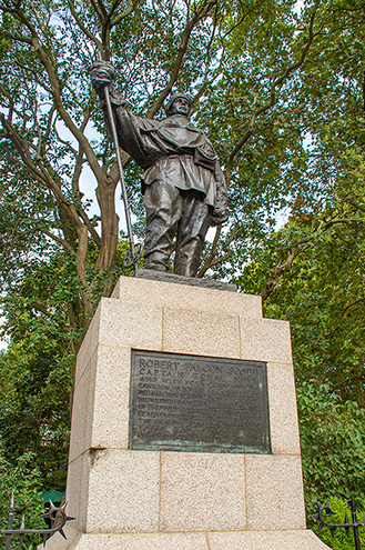 The image shows a statue of a man holding a torch, standing on a pedestal with a plaque. The statue is situated in an outdoor setting, surrounded by trees. The plaque on the pedestal contains text, likely providing information about the statue or the person it represents.