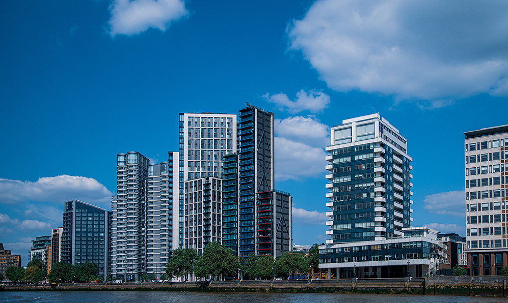 The image depicts a row of modern high-rise buildings along a waterfront, under a clear blue sky with scattered clouds. The buildings vary in architectural design, featuring a mix of glass and concrete facades. Trees and a pedestrian walkway are visible along the water's edge, suggesting an urban waterfront development.