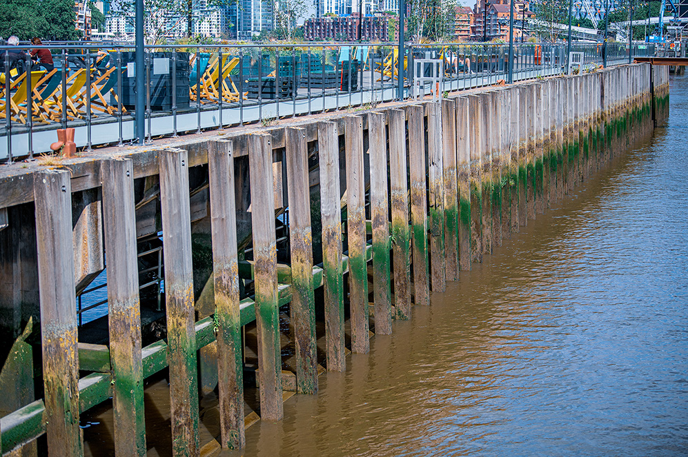 The image depicts a waterfront scene featuring a wooden pier or walkway extending along a body of water. The wooden structure shows signs of wear and algae growth, indicating it has been in place for some time. The pier is equipped with railings for safety, and there are various structures and buildings visible in the background, suggesting an urban setting. The water appears calm, and the overall scene suggests a mix of natural and man-made elements in a city environment.