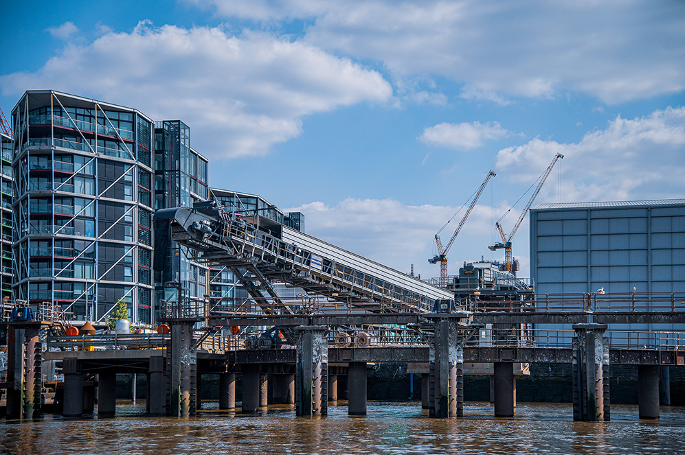 The image depicts an industrial waterfront facility with a large warehouse-like structure. There are cranes in the background, and a barge is docked at the loading platform, which has various equipment and safety features such as railings and ladders. The facility appears to be involved in shipping or manufacturing activities.