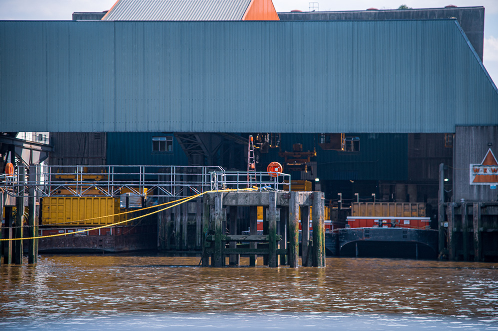 The image shows a waterfront industrial area with a large, weathered building and a dock. The dock has railings and is equipped with various industrial elements such as containers and machinery. The water appears to be calm, and there are signs of wear and tear on the structures, indicating a well-used facility.