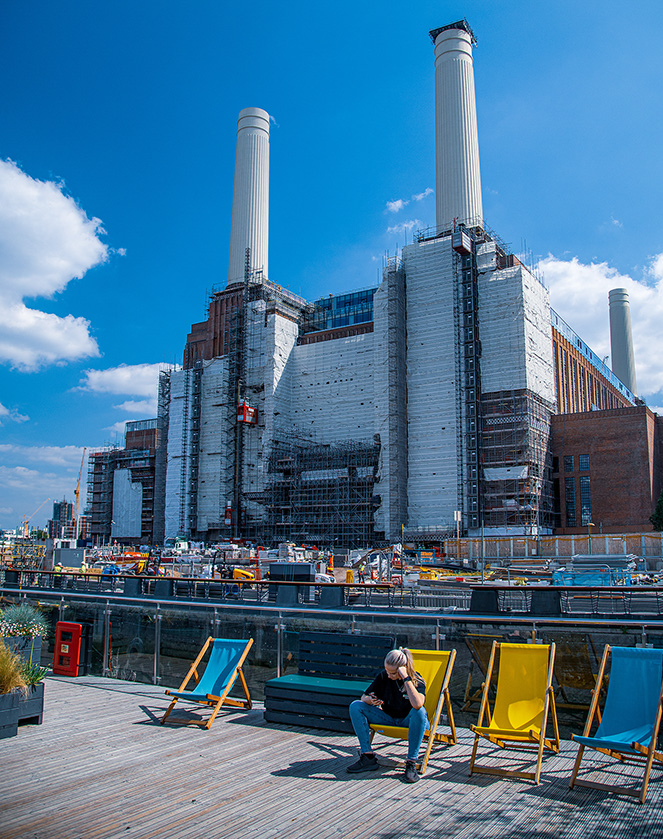he image depicts a large industrial building with multiple smokestacks under construction or renovation. In the foreground, there is a wooden deck area with colorful deck chairs and a person sitting on one of the chairs, facing away from the camera. The scene is set on a clear, sunny day with a bright blue sky and some clouds.