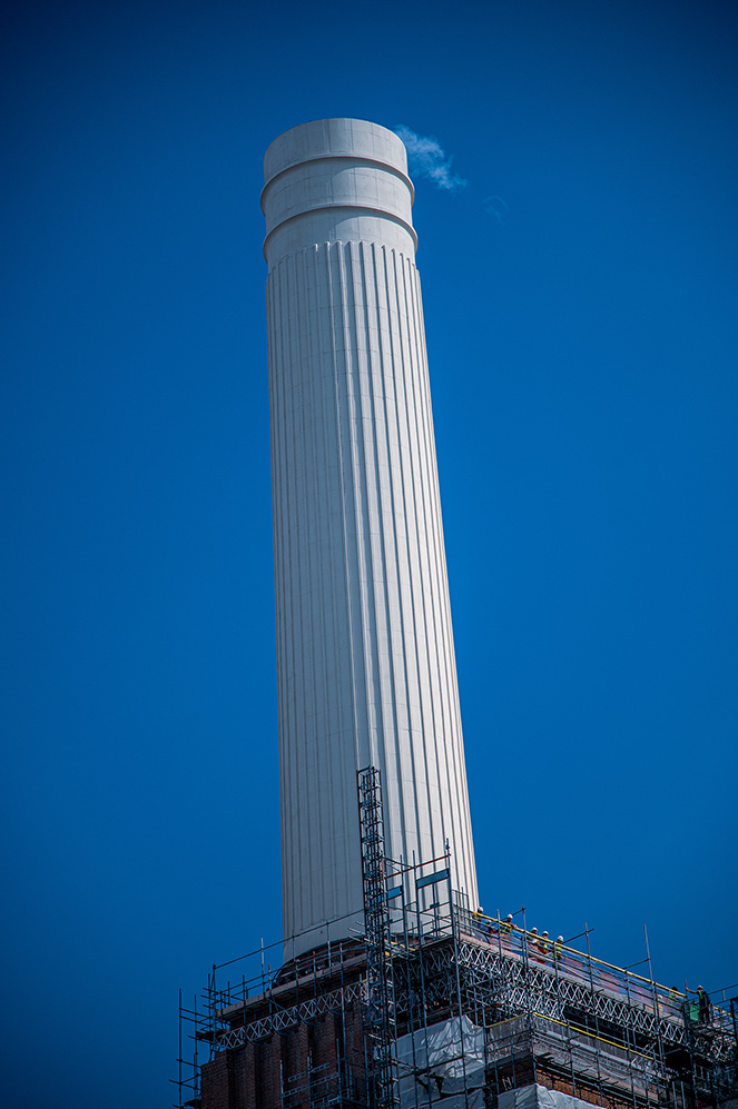 The image shows a tall, cylindrical chimney or smokestack with a white exterior and vertical grooves. The structure is under construction or renovation, as evidenced by the scaffolding surrounding its base. The sky in the background is clear and blue, with a small amount of smoke or steam emanating from the top of the chimney.