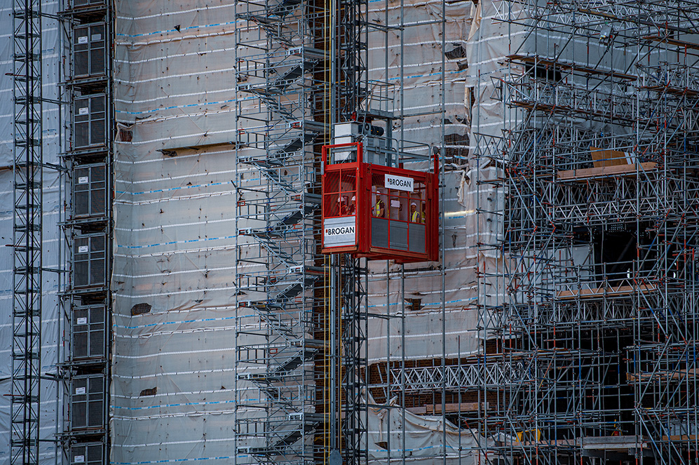 The image depicts a construction site with scaffolding covering a building. A red lift with the brand name 'PROGAN' is visible, carrying workers. The building appears to be in the process of renovation or construction, with protective coverings and various construction materials present.