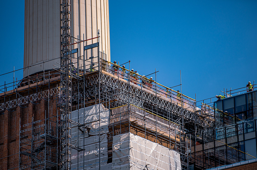 The image shows a construction site with scaffolding erected around a tall building. Workers in safety gear are visible on the scaffolding, indicating ongoing maintenance or renovation work. The building appears to be a significant structure, possibly a historical or important landmark, given its size and architectural details.