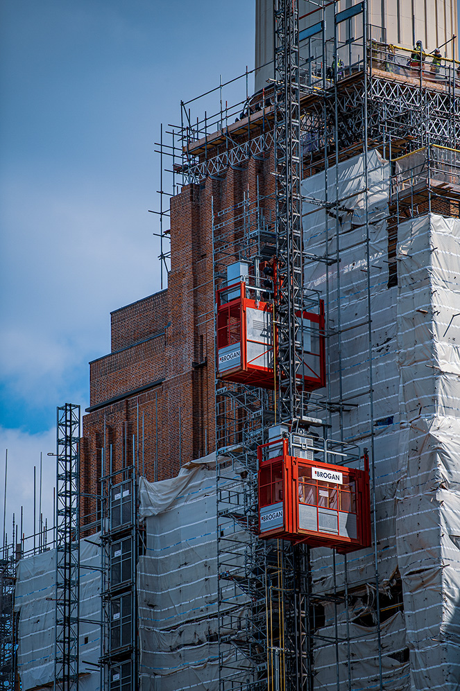 The image shows a building under construction or renovation, covered in scaffolding and protective white netting. Two red elevators, labeled 'BROGAN,' are visible, transporting workers and materials up and down the structure. The sky is clear with a few clouds, indicating good weather conditions for construction work. The building appears to be multi-storied, with a mix of brick and modern glass elements.
