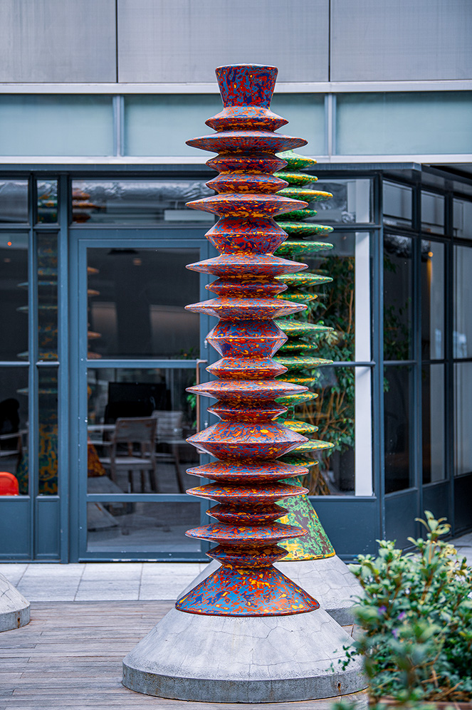 The image depicts a tall, colorful sculpture composed of multiple stacked, disc-like elements. The sculpture is situated outdoors, in front of a modern building with large glass windows. The base of the sculpture is wide and appears to be made of stone, while the stacked discs are vibrant and multicolored, creating a visually striking contrast against the more muted surroundings. The area around the sculpture includes some greenery and a wooden deck.