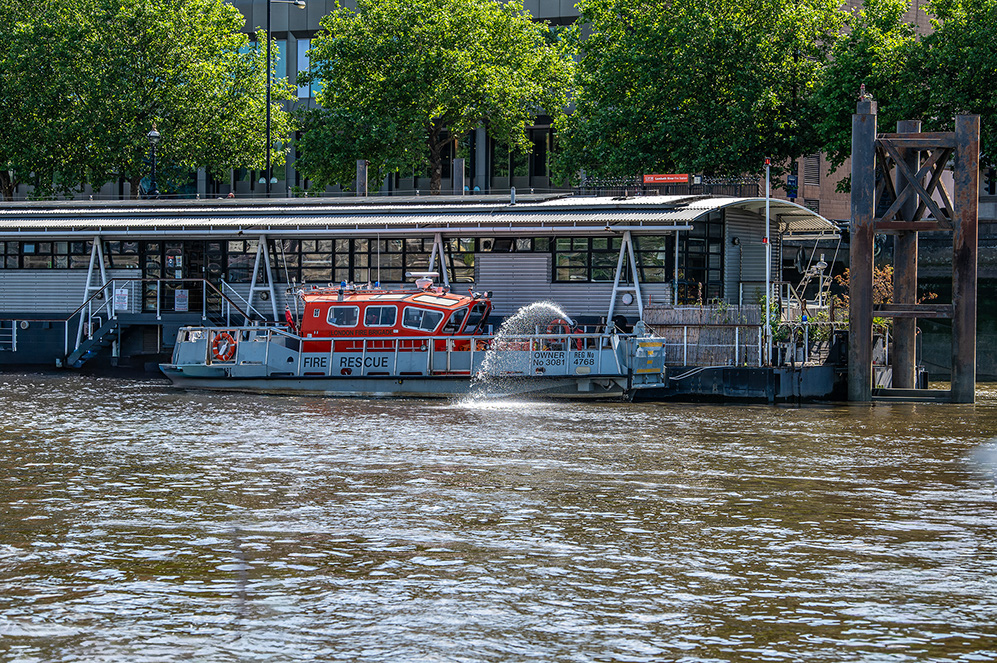 The image shows a fire rescue boat docked at a pier. The boat is marked with 'FIRE RESCUE' and has the owner's name and registration number displayed. The pier has a small building with a corrugated metal roof and is situated along a river with trees and buildings in the background.>
                                    <figcaption>
                                         <meta itemprop=