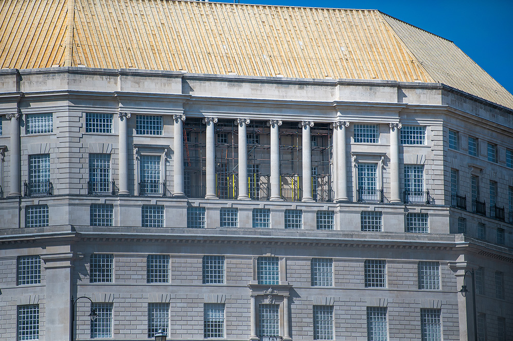 The image depicts a section of a grand, classical-style building with a curved facade. The building features multiple windows with wrought iron railings and is adorned with columns and decorative stonework. The roof appears to be made of a golden, metallic material, contrasting with the grey stone of the building. The architectural style suggests it could be a historic or significant public building, such as a government or institutional structure.