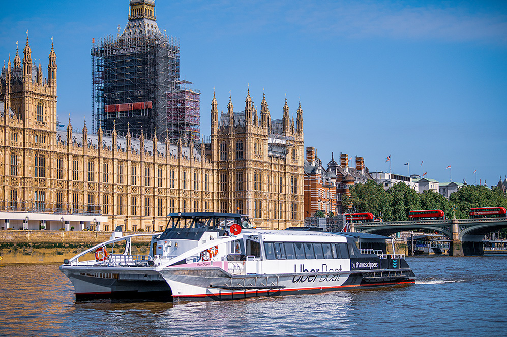 The image shows a river scene with a modern boat labeled 'Uber Boat by Thames Clippers' in the foreground. In the background, there is a large, historic building under construction, likely the Palace of Westminster, with scaffolding and cranes visible. The sky is clear, and there are other structures and greenery along the riverbank. A bridge with red double-decker buses is also visible in the background.