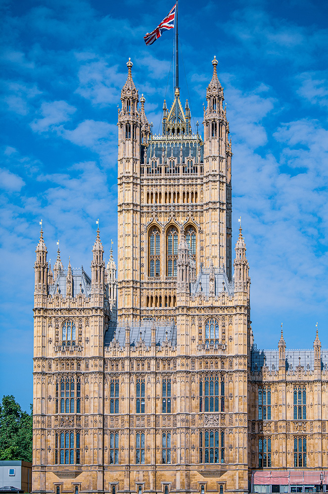 The image depicts the Palace of Westminster in London, United Kingdom, with the Union Jack flying atop the Victoria Tower. The Gothic architecture is prominently featured, showcasing intricate details and multiple spires against a backdrop of a clear blue sky with scattered clouds.