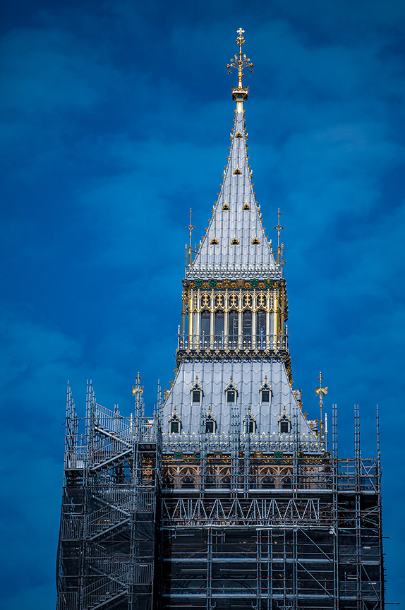 The image shows a tall, ornate spire under construction or renovation. The spire is adorned with intricate designs and gold accents, and is topped with a cross. Scaffolding surrounds the lower portion of the spire, indicating ongoing work. The sky in the background is clear and blue, providing a stark contrast to the detailed structure.
