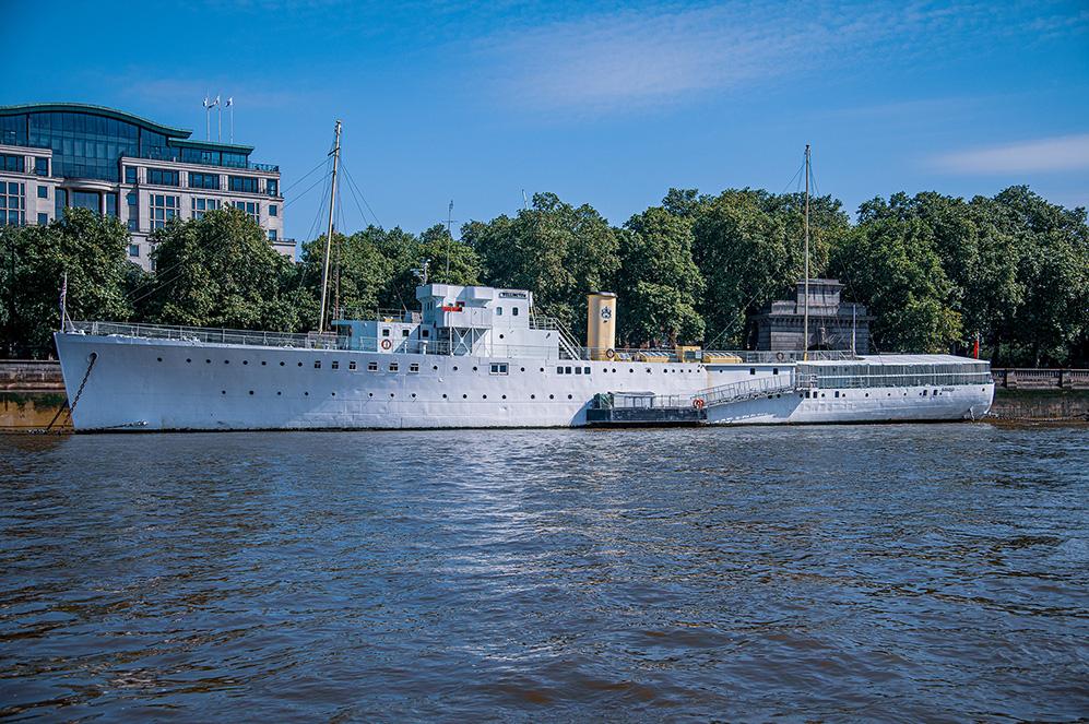 The image shows a large, white ship docked on a river, with modern buildings and lush green trees in the background under a clear blue sky.