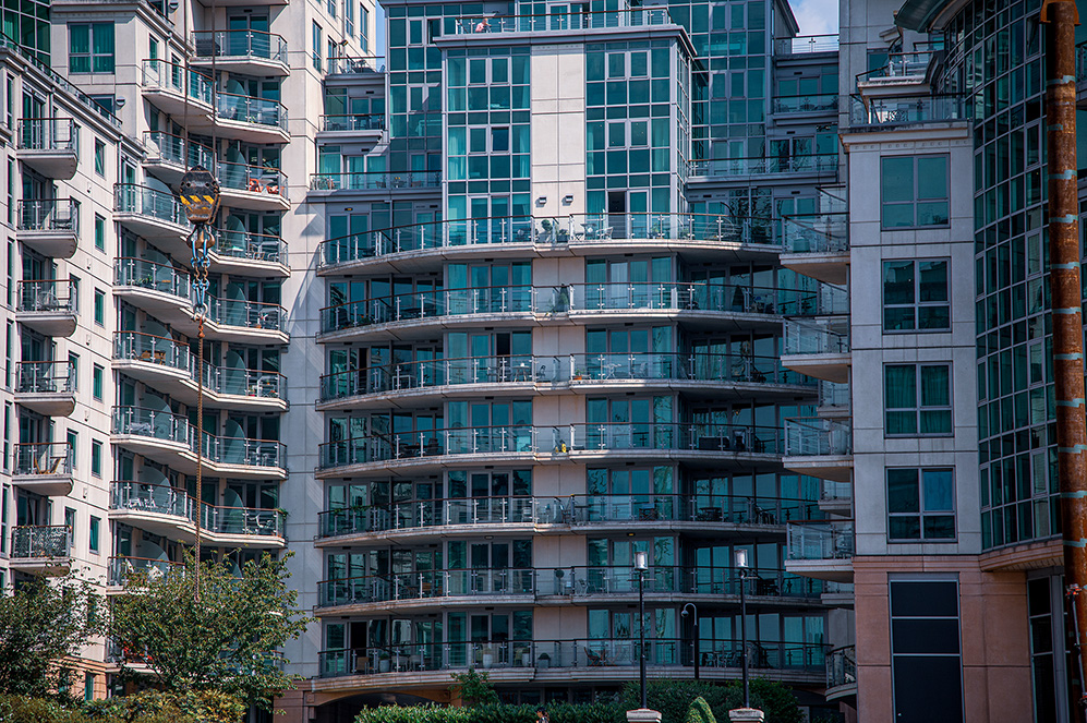 The image depicts a modern multi-story residential building with numerous balconies, large windows, and a mix of glass and concrete facade. The balconies are curved and have glass railings, and the building appears to be well-maintained with some greenery visible at the base.