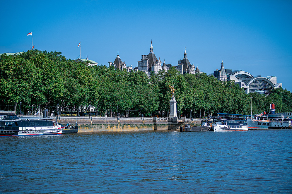 The image depicts a scenic view of a river with boats, including an Uber Boat, and a historic building with spires in the background. The building is partially obscured by a line of lush green trees. The sky is clear and blue, suggesting a bright, sunny day. The scene captures a blend of natural and architectural beauty, likely in a city setting.